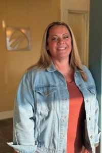 Dental staff member Rachel wearing denim jacket over coral top, smiling in office lobby with logo sign.